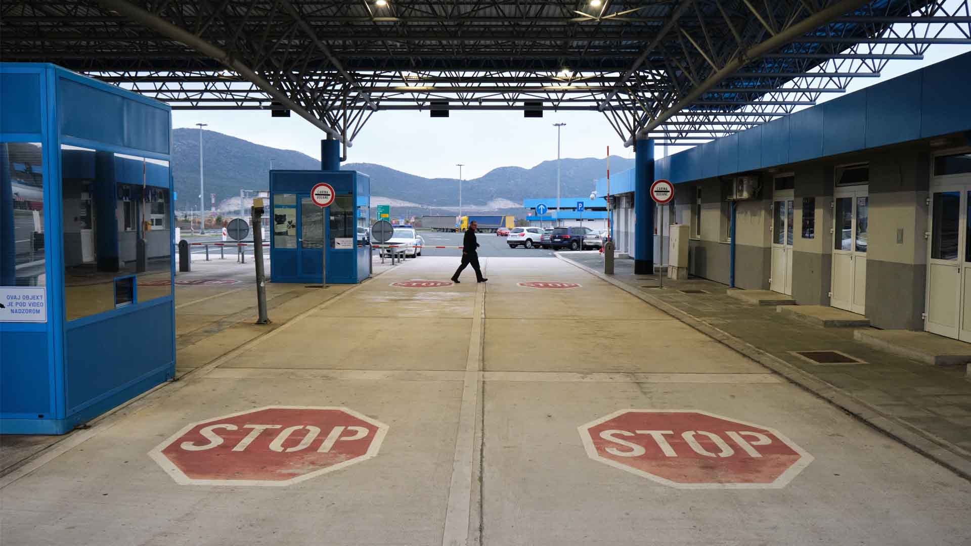 This is a picture of the Croatian customs on the border with Bosnia and Herzegovina. We see the pavement in the foreground of the picture. Two large STOP signs are painted on the ground. Further down we see two boom barriers/traffic barriers. A person walks through the center of the frame in front of the barriers.