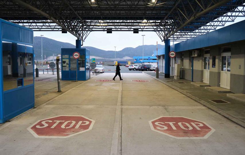This is a picture of the Croatian customs on the border with Bosnia and Herzegovina. We see the pavement in the foreground of the picture. Two large STOP signs are painted on the ground. Further down we see two boom barriers/traffic barriers. A person walks through the center of the frame in front of the barriers.