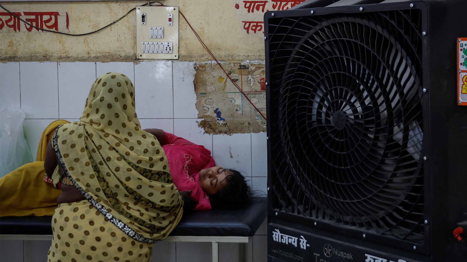 A relative stands next to a patient lying on a strecher inside an emergency ward at a hospital in Ballia District in the northern state of Uttar Pradesh, India, June 21, 2023. Beside them is a large black fan.