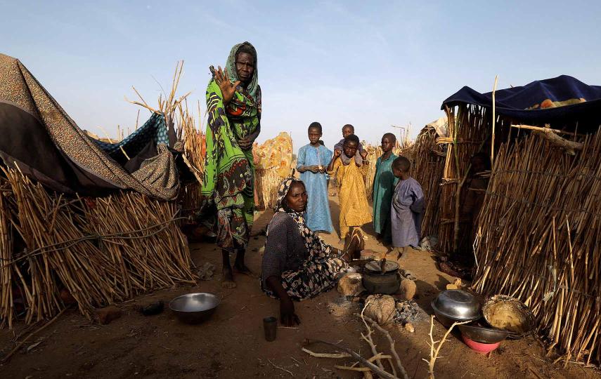 Ashta, a Sudanese refugee, who has fled the violence in her country, reacts as she walks past her relatives and makeshift shelters, near the border between Sudan and Chad, in Koufroun, Chad, May 9. Around Ashta are a woman sitting on the ground and five children behind her. They are in betweeen makeshift homes made of wooden sticks.