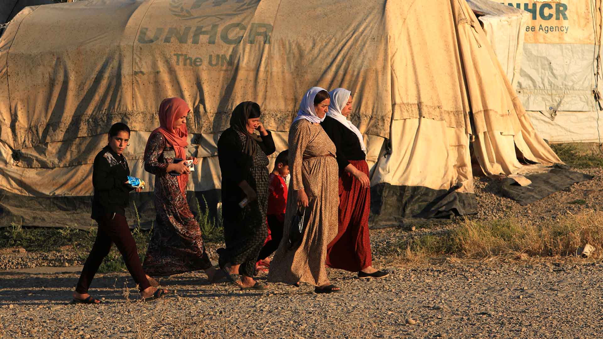 Five women and two children are pictures walking through Khanke Camp. Behind them we see tents all labeled with the UNHCR logo.