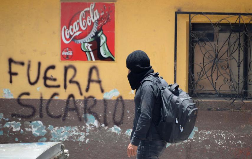 A member of the new self-defense group known as 'El Machete' walks by a graffiti reading 'Sicario out' as he looks for members of drug gangs and municipal authorities during a protest against the growing violence in the area, in Pantelho, in Chiapas state, Mexico. He wears a face covering, jeans, a dark jumper and a back pack. The photo was taken on July 27, 2021.