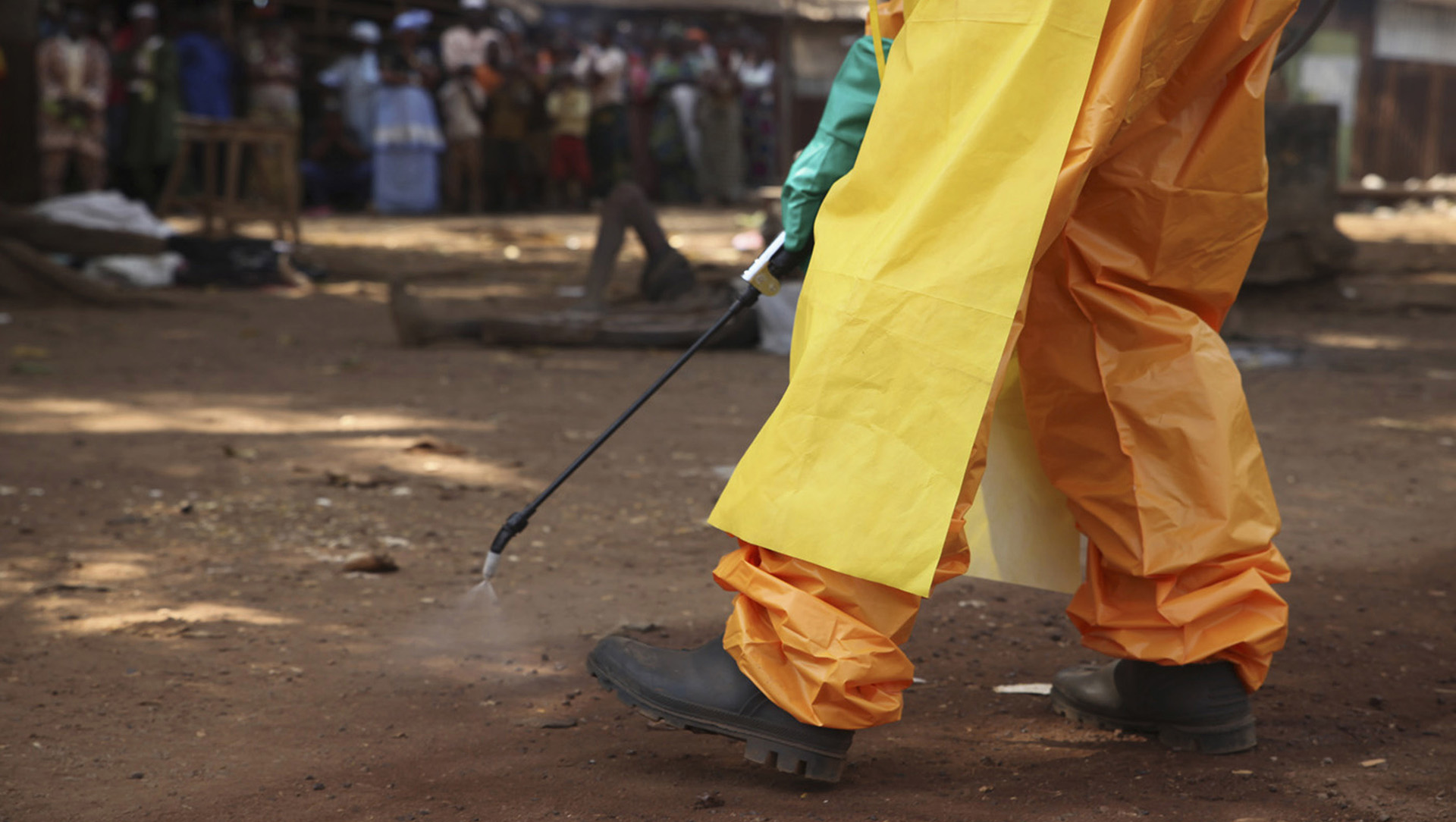 The image shows the bottom half of a person's body - dressed in a hazmat suit, apron, gloves, and boots - holding a disinfectant wand spraying the ground. 