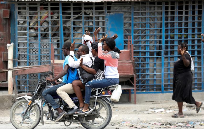 Five residents of Port-au-Prince Haiti can be seen in the photo. Four are on a motorcycle and one walking closely behind it. All except the motorcycle driver has their hands up in the air. They are fleeing gang violence.
