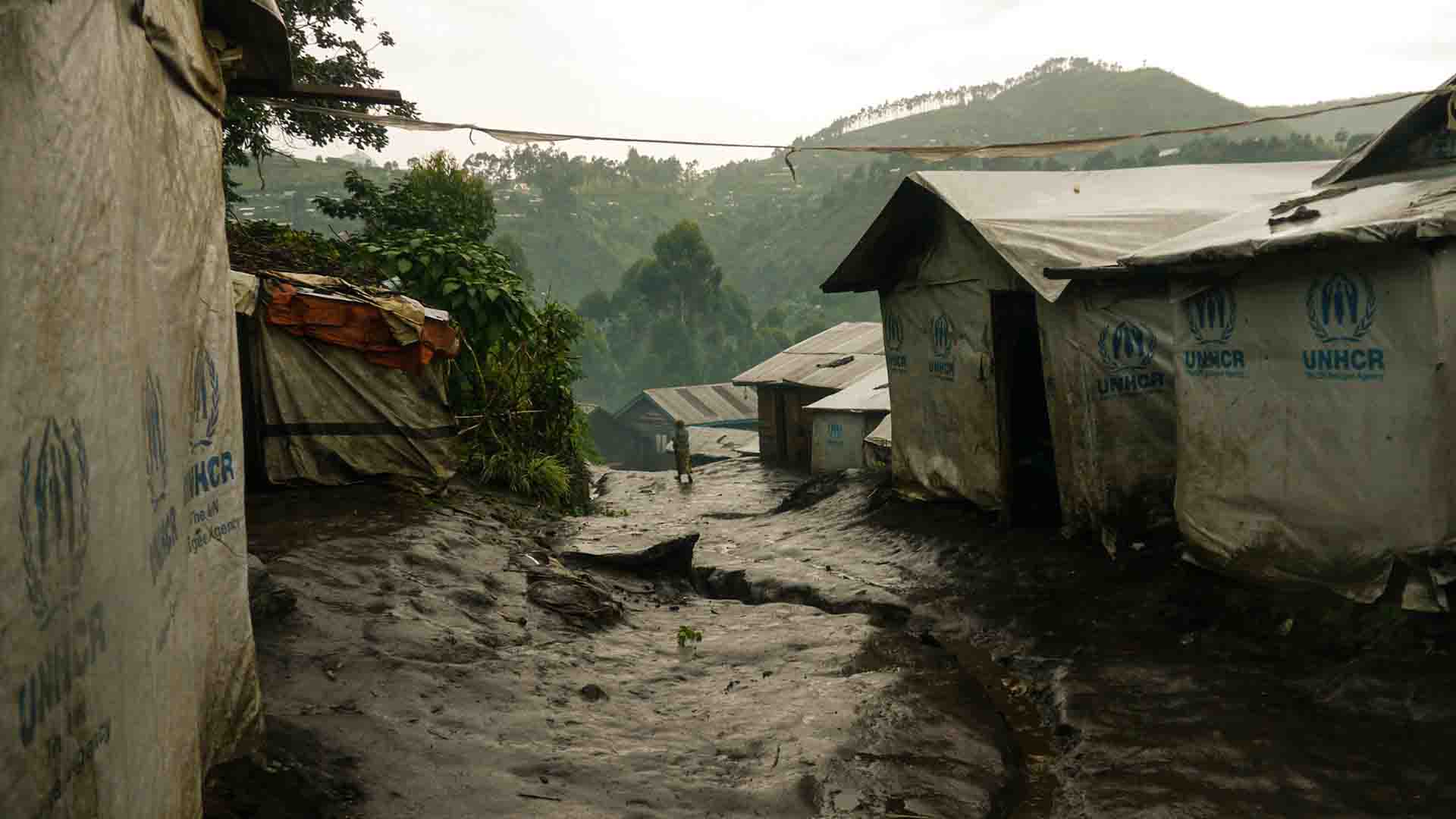 This is a picture of Kalinga Camp located in North Kivu in the DRC. It looks like it recently rained, the dirt street looks damp. Houses on either side of the road have tarps with UNHCR logos covering them.
