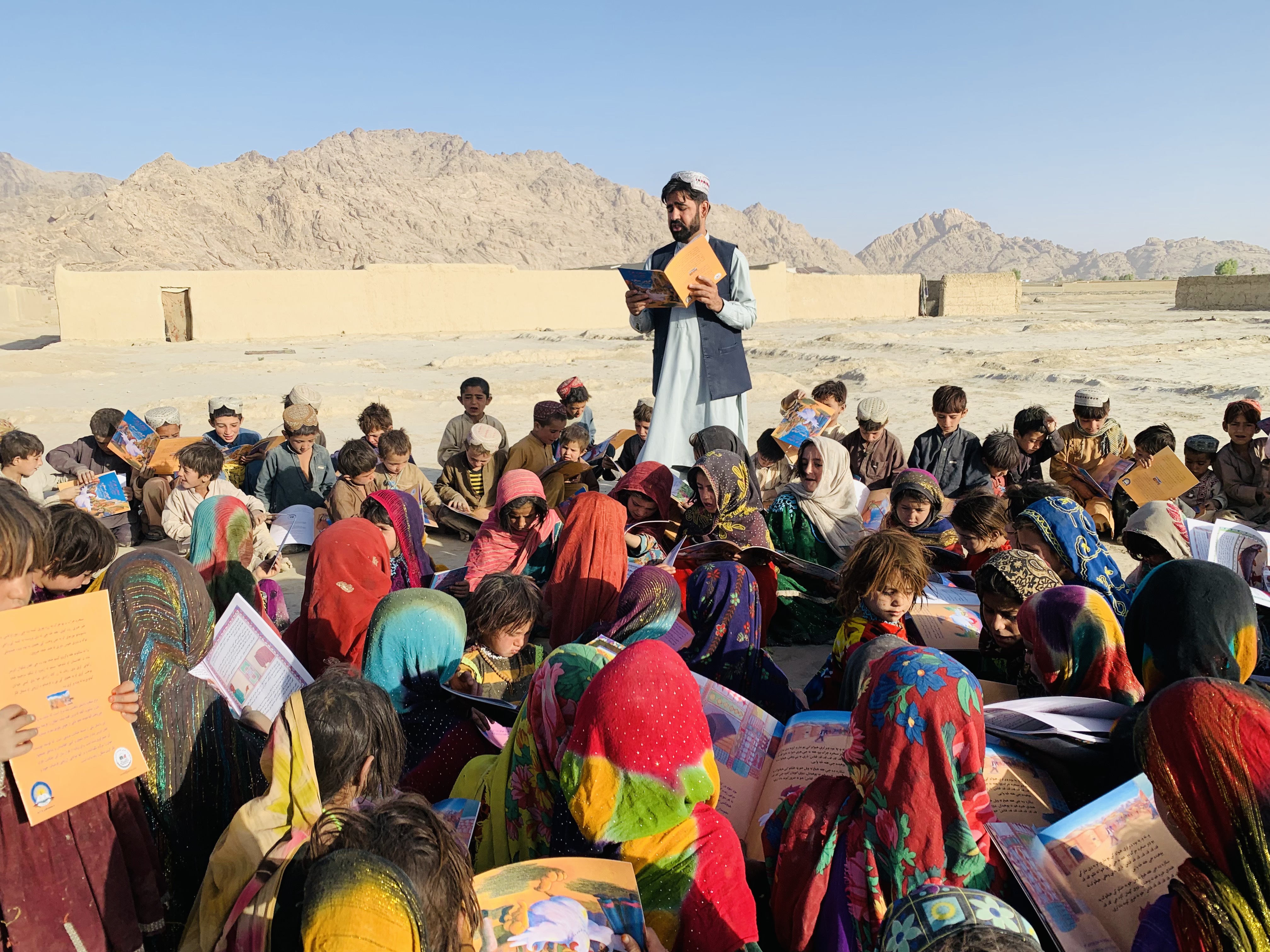 A man stands and reads a book to dozens of children who are sitting in the ground. 