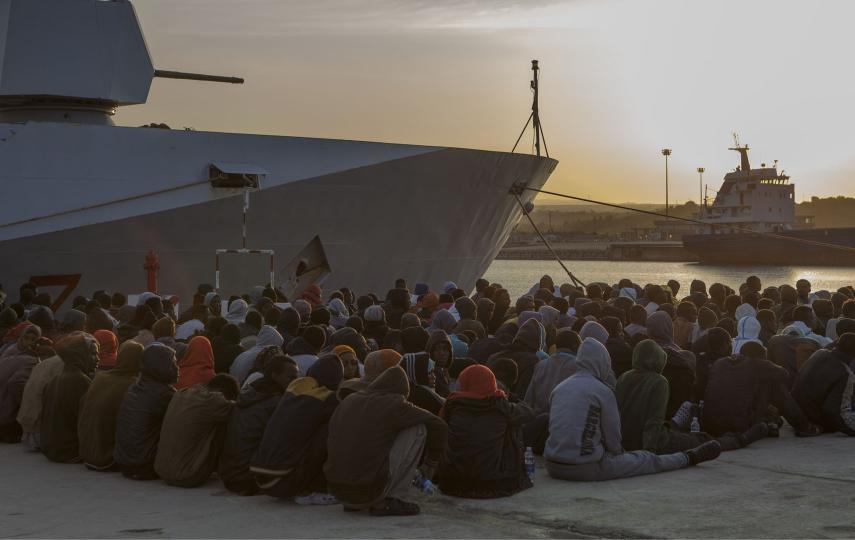 Pictured are around 400 African asylum seekers and migrants wait after disembarking in the Italian port of Augusta, Sicily. They sit on the ground, in the background the sun is setting and to the right we see a ship. 