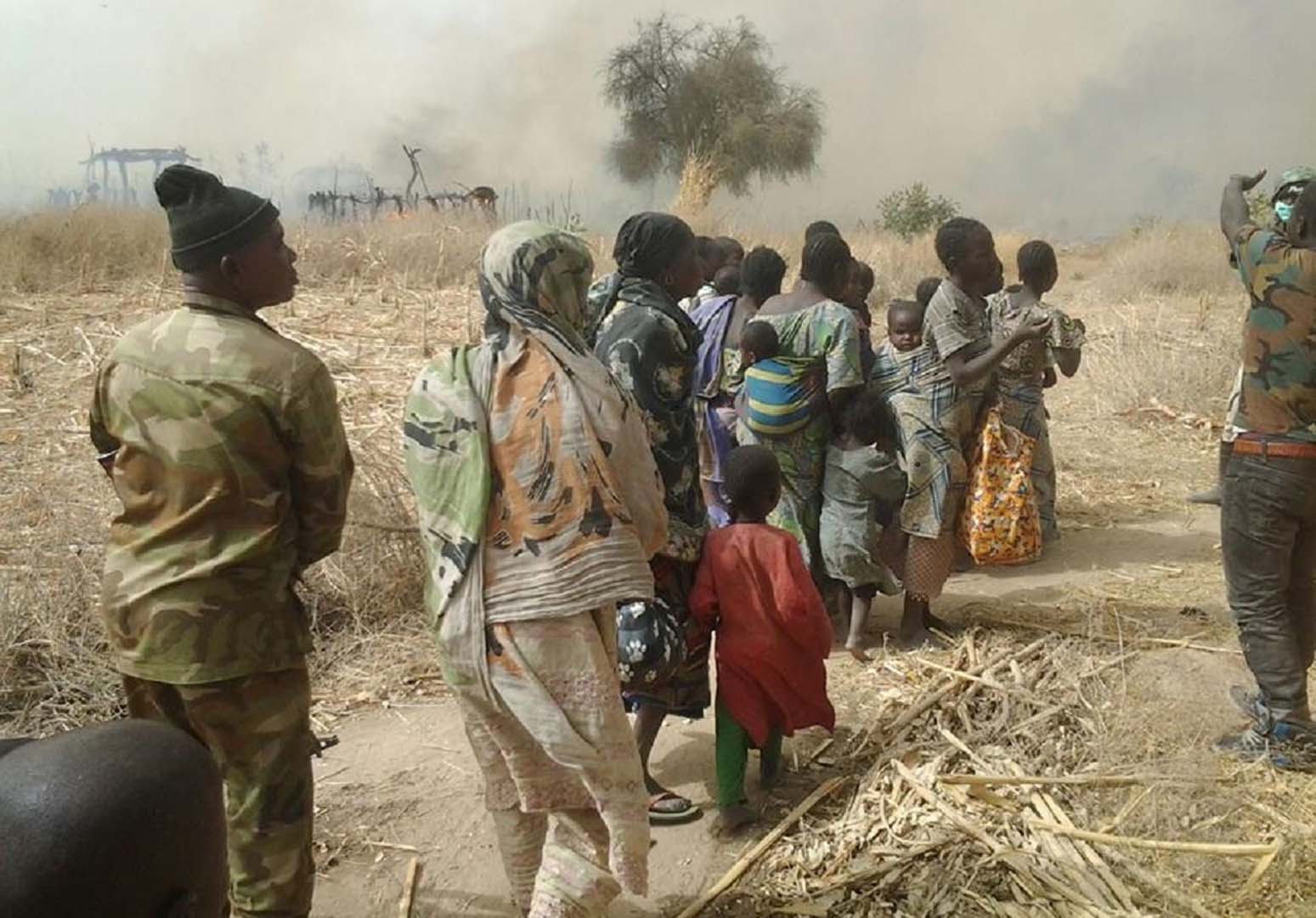 A group of people stand in line, two apparent military men are around them. In the distance we see a village burning. 