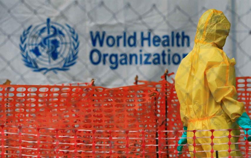 Person with hazmat suit walking in front of a WHO logo. 