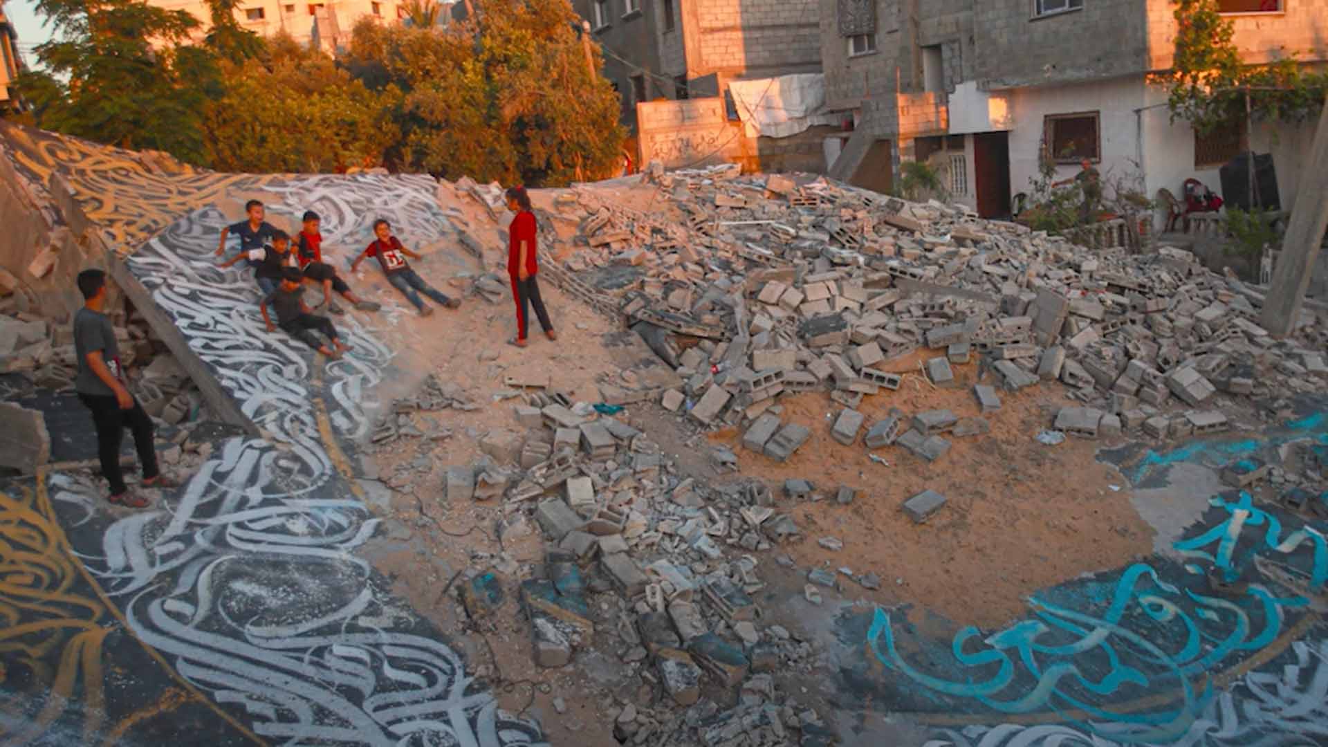 A group of young children lie on the ground with rubble around them, parts of the floor have been covered in art.