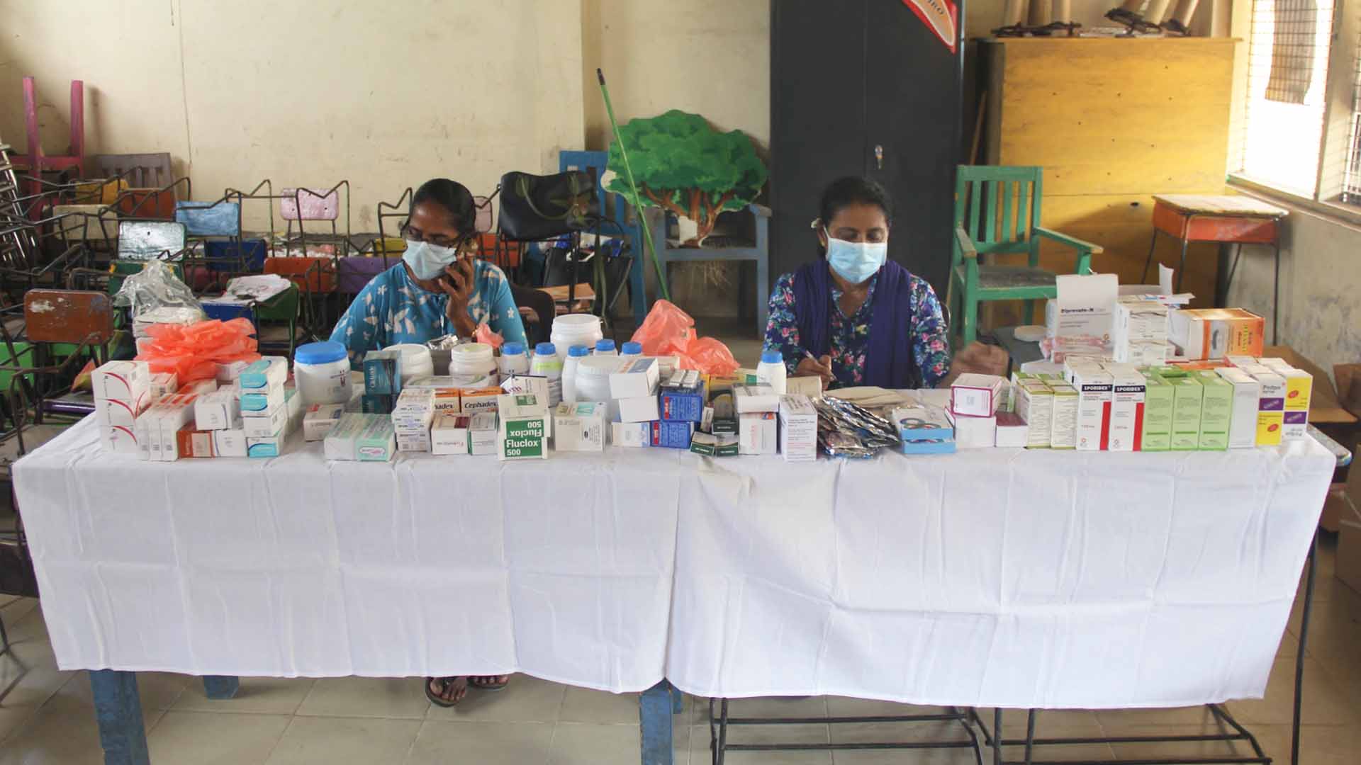The picture shows a table covered with a white cloth. The table has many boxes of medicine. Two women wearing surgical masks sit behind the table.