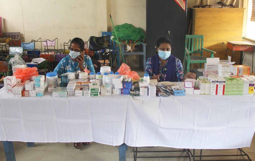The picture shows a table covered with a white cloth. The table has many boxes of medicine. Two women wearing surgical masks sit behind the table.
