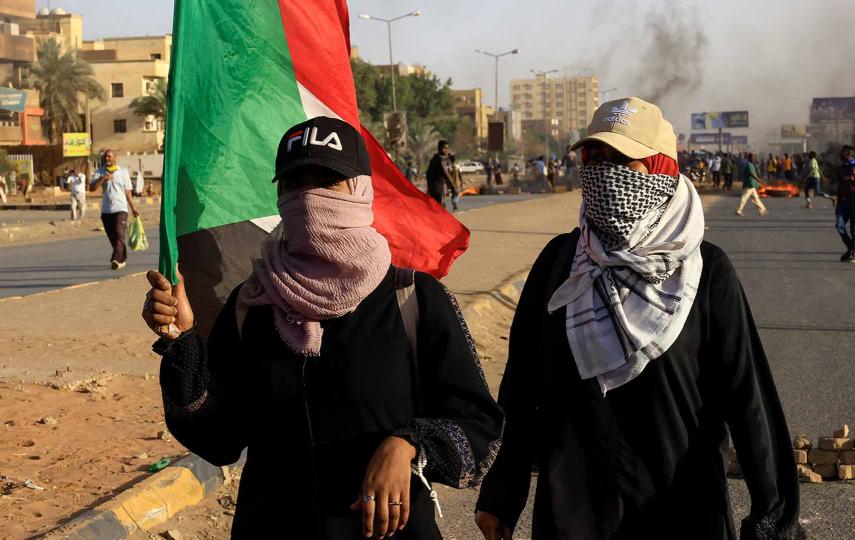 In this photo, we see two young women. Their faces are covered and they're both wearing caps. One of them holds the flag of Sudan in one hand. In the background we see smoke and dispersed people also protesting.
