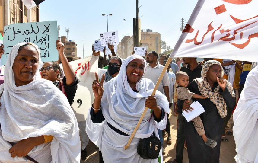Pictured are women members of the Sudanese Women’s Union attend a rally in December 2022 in Khartoum. They're wearing white robes and head coverings. At the centre is a woman. She is smiling and holding out a peace sign with her right hand while holding a large flag with the left.