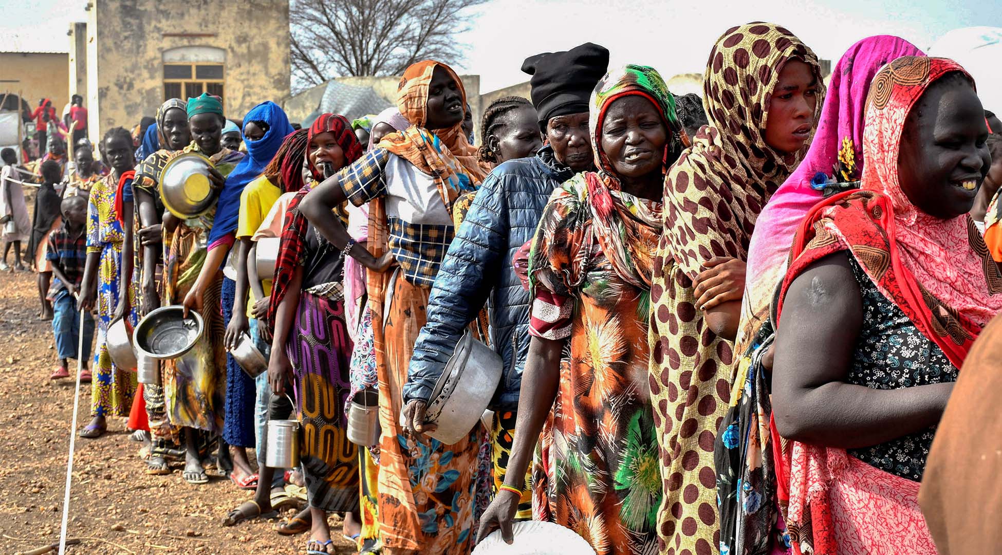 Pictured are many women standing in line. Some of them hold metal pots and plates. They wear ankle-length robes of different colors. The day seems sunny. 