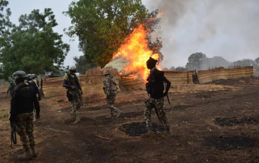 A group of soldiers stand in the foreground as a fire blazes behind the,.